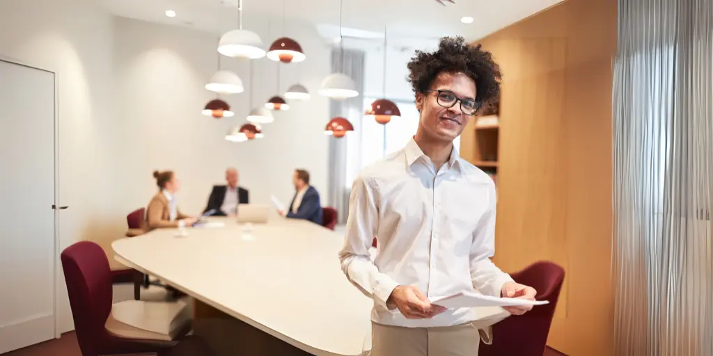 black young man in office smiling and holding papers with other office workers behind him